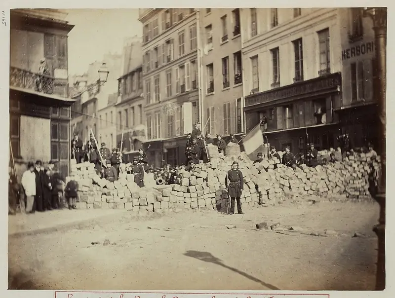 Old sepia photograph of a waist-high barricade of stacked paving stones across a Paris street, with people standing on and around it.
