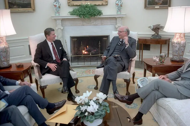 Photograph of President Reagan and Fed Chair Paul Volcker seated in armchairs talking by the Oval Office fireplace, with other officials seated in the foreground.