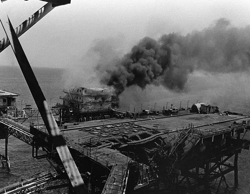 Black-and-white photo of an offshore oil platform with a wrecked deck and a building pouring thick black smoke, seen through a steel railing.