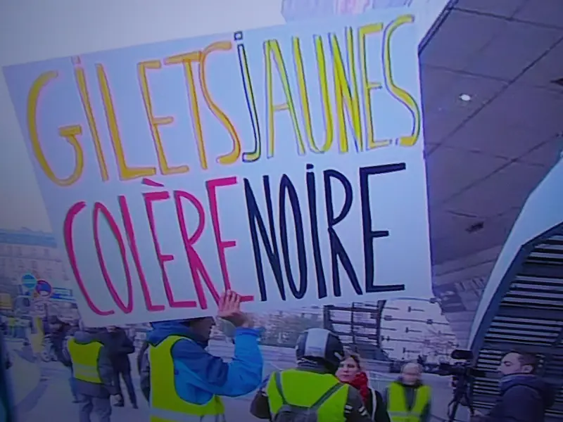 Protester holding up a hand-lettered sign reading "Gilets jaunes, colère noire" above people in high-visibility yellow vests.