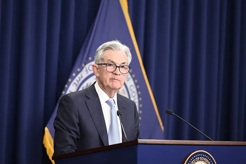 Federal Reserve Chair Jerome Powell, wearing glasses and a dark suit, speaks at a podium in front of a blue curtain and flags.